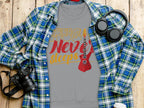 On a wooden surface, the Garment Graphics vibrant gray t-shirt features a red electric guitar and Music Never Sleeps. Ideal for music lovers, it pairs well with the blue and green plaid shirt, headphones, camera, and white cup.