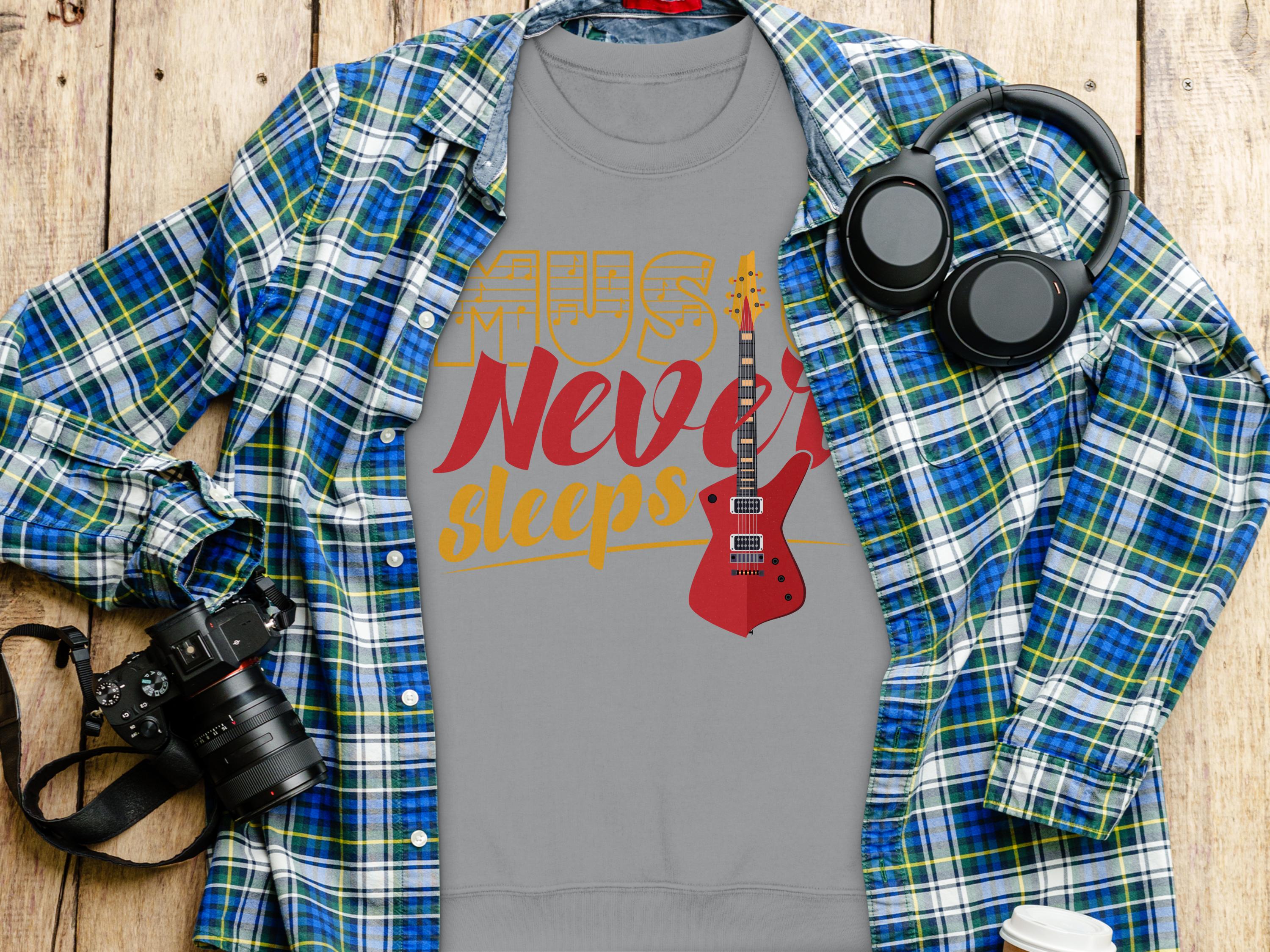 On a wooden surface, the Garment Graphics vibrant gray t-shirt features a red electric guitar and Music Never Sleeps. Ideal for music lovers, it pairs well with the blue and green plaid shirt, headphones, camera, and white cup.