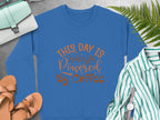 A blue graphic sweatshirt from Garment Graphics, featuring This day is powered by coffee in vibrant orange text, rests on a concrete surface. Its playfully styled with tan sandals, a striped shirt, a mint handbag, a wristwatch, and green fern leaves.
