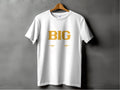 A Garment Graphics classic fit white t-shirt, hung on a wooden hanger against a dark background, features BIG in large yellow letters and subtle humorous designs underneath.