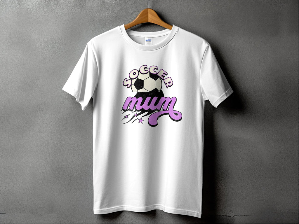 A white T-shirt from Garment Graphics features Soccer Mum in stylized text with a dynamic soccer ball, lines, and stars. This classic fit tee is expertly designed and gracefully hangs on a wooden hanger against a gray background.