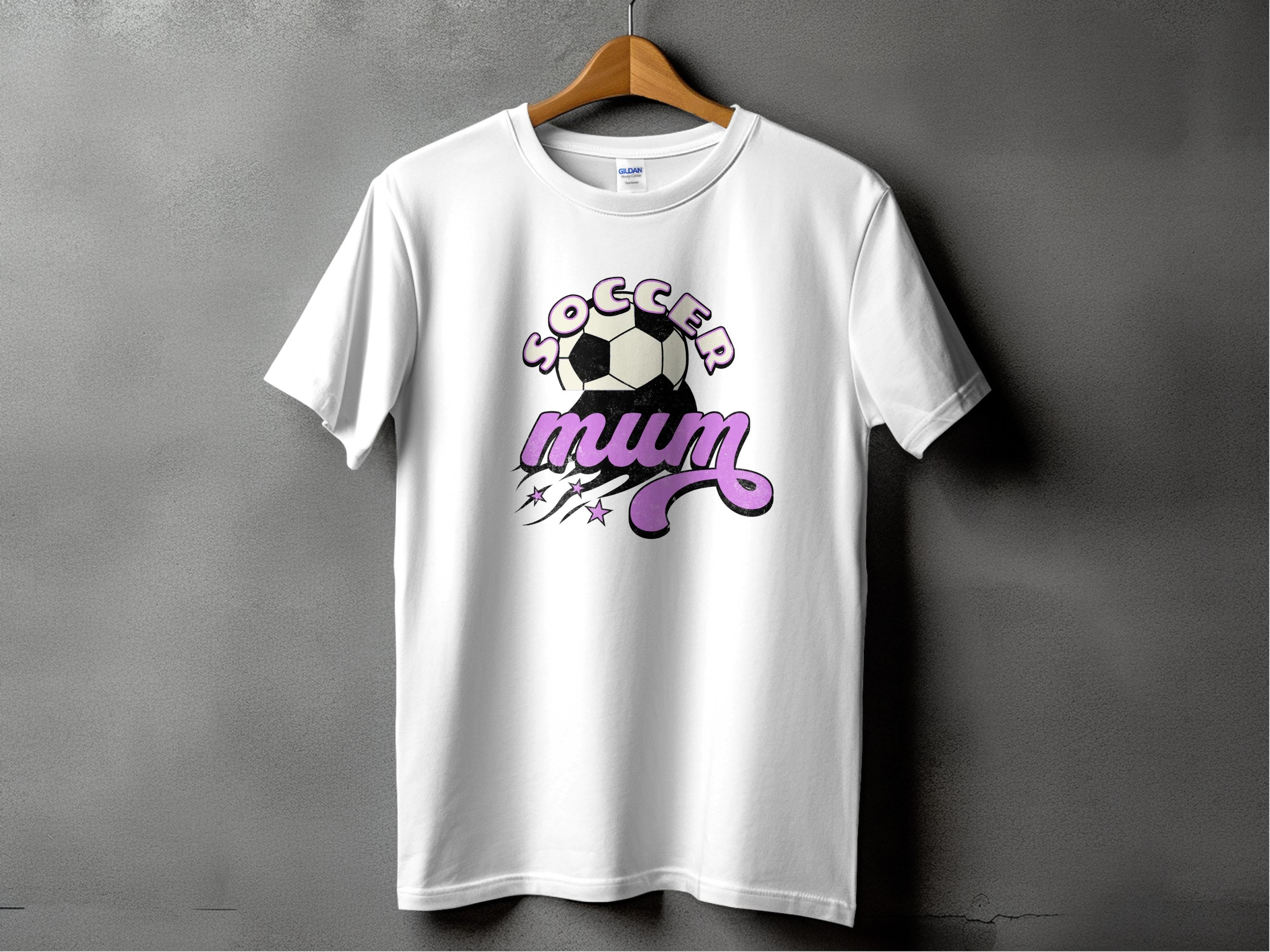 A white T-shirt from Garment Graphics features Soccer Mum in stylized text with a dynamic soccer ball, lines, and stars. This classic fit tee is expertly designed and gracefully hangs on a wooden hanger against a gray background.