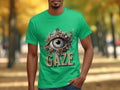 Someone in a Garment Graphics classic fit green T-shirt showcases a detailed eye illustration with ornate patterns. Below it, the word GAZE is prominently displayed. A blurred outdoor scene of trees and a path enhances the shirts unique design allure.