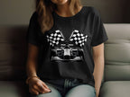 A motorsport enthusiast with long hair sits on a couch, wearing a black Garment Graphics t-shirt featuring a race car and crossed checkered flags, complemented by a stylish bracelet on their wrist.