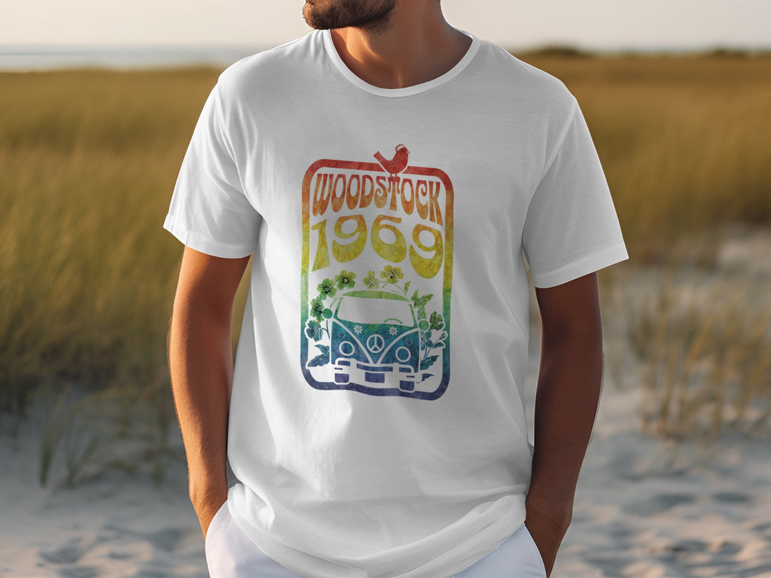 On a sandy beach, a music lover wears a Garment Graphics white T-shirt featuring Woodstock 1969 and a colorful retro van design. Tall grass sways gently in the breeze beneath the clear sky.