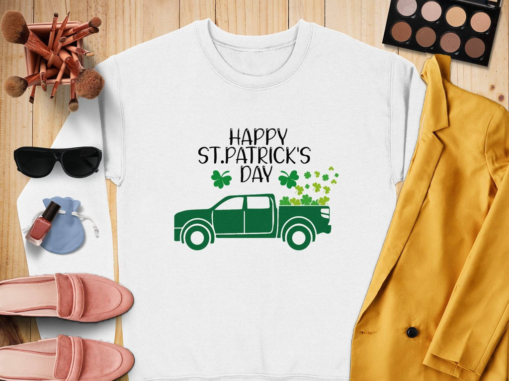 A flat lay displays a festive Garment Graphics St. Patricks Day sweatshirt with a green truck design, surrounded by makeup brushes, an eyeshadow palette, sunglasses, a light blue pouch, pink loafers, nail polish, and a mustard yellow jacket for a cheerful look.