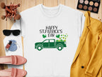 A flat lay displays a festive Garment Graphics St. Patricks Day sweatshirt with a green truck design, surrounded by makeup brushes, an eyeshadow palette, sunglasses, a light blue pouch, pink loafers, nail polish, and a mustard yellow jacket for a cheerful look.