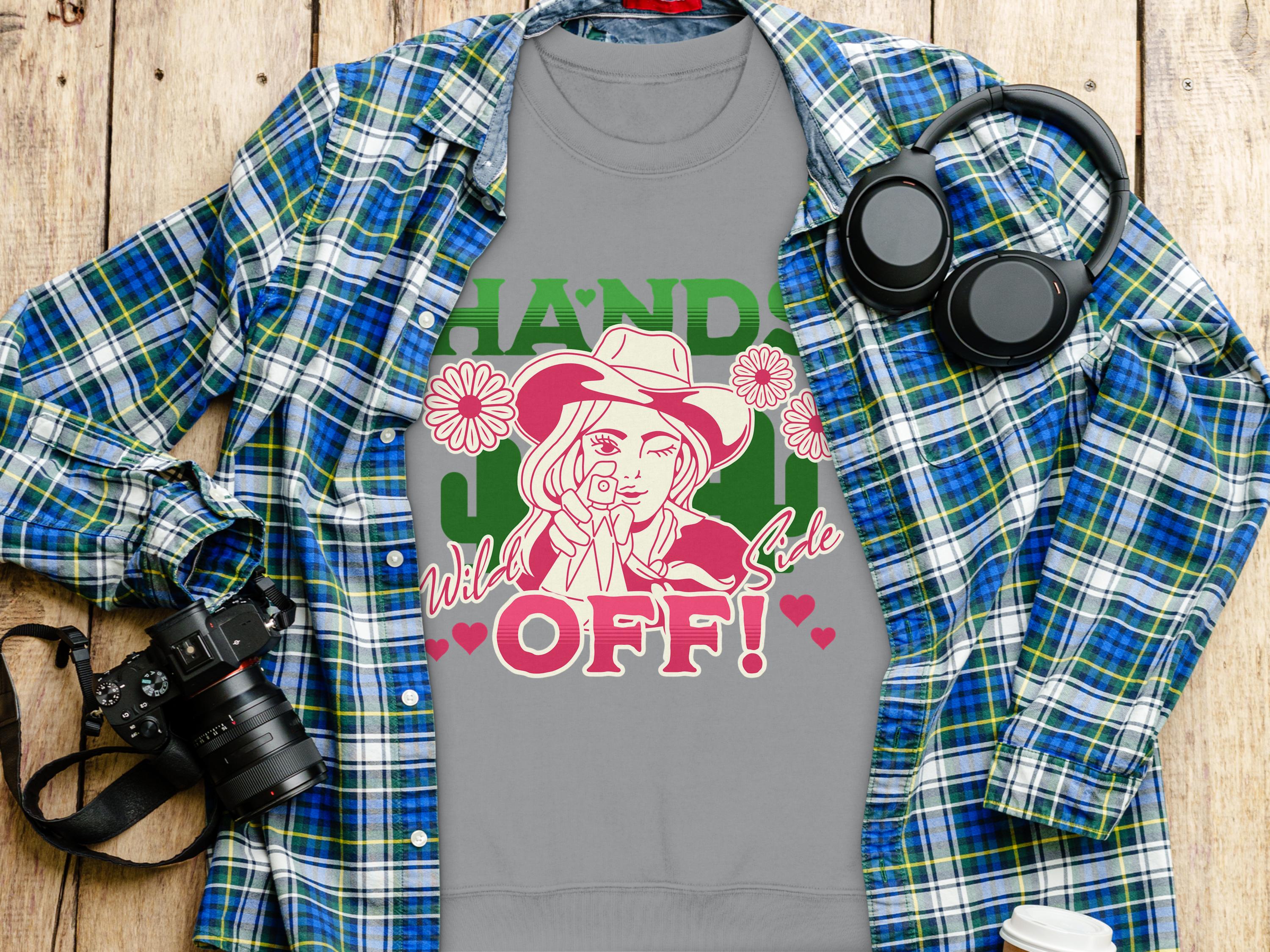 A flat lay image features a blue and green plaid shirt under a Hands Off! graphic tee by Garment Graphics, surrounded by items like a camera, headphones, and a loose fit takeaway coffee cup on the wooden surface.