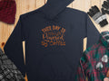 Garment Graphics offers a stylish dark blue hoodie with This Day Is Powered by Coffee in brown script. Ideal for coffee lovers, its surrounded by a knitted hat, striped gloves, and plaid fabrics on a wooden surface.