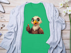 A green Garment Graphics Eagle T-shirt with a cartoon eagle giving a thumbs up is laid on light wood, paired with a gray cardigan, surrounded by white flowers, perfume, and classic fit white pointed shoes—ideal for a fun casual wardrobe.