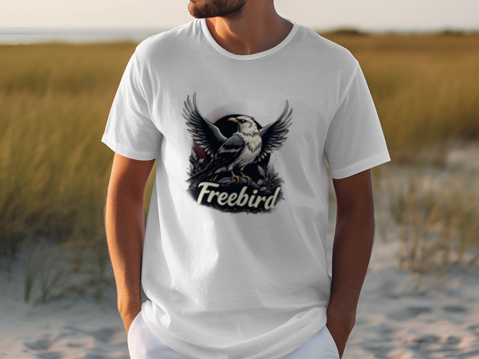 An individual sporting a bold graphic Freebird eagle T-shirt by Garment Graphics, crafted from 100% cotton, stands outdoors with blurred grass and sand as the backdrop, highlighting their style.