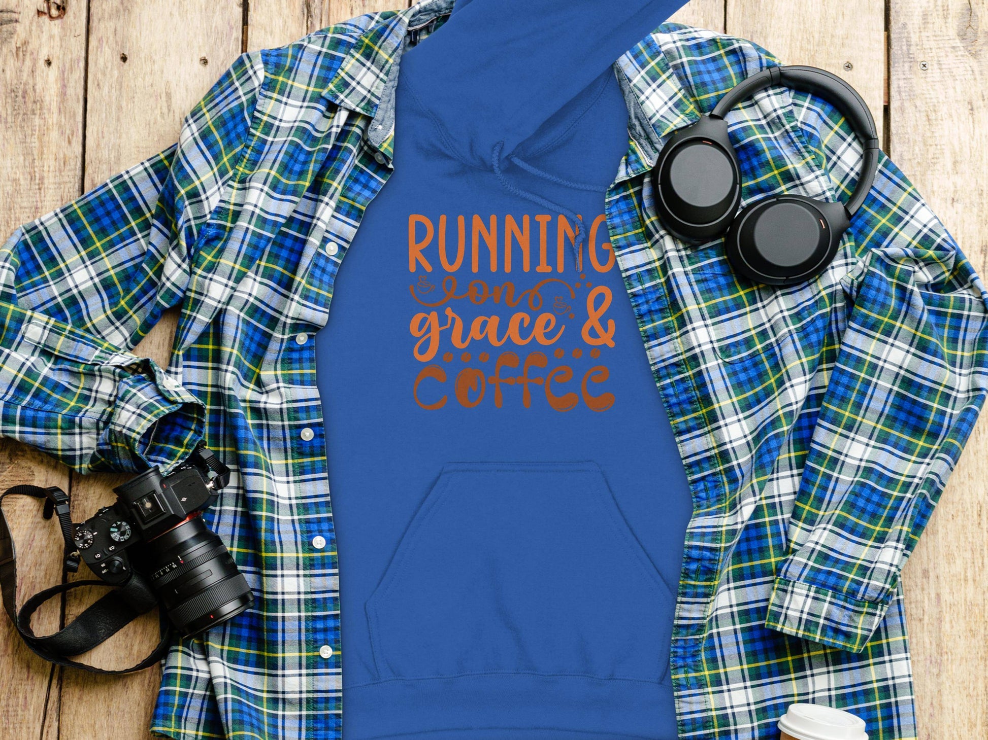 A blue medium-heavy fabric hoodie by Garment Graphics, featuring Running on Grace & Coffee in orange, lies on a wooden surface. This classic fit is paired with a blue plaid shirt. A camera, headphones, and a white disposable cup are nearby.