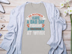 Displayed on a wooden surface, this Garment Graphics gray T-shirt, featuring Id rather have a bad day fishing than a good day at work, makes an ideal gift for anglers. Its partly covered by a light gray cardigan and accompanied by a flower, wristwatch, perfume, and nail polish.