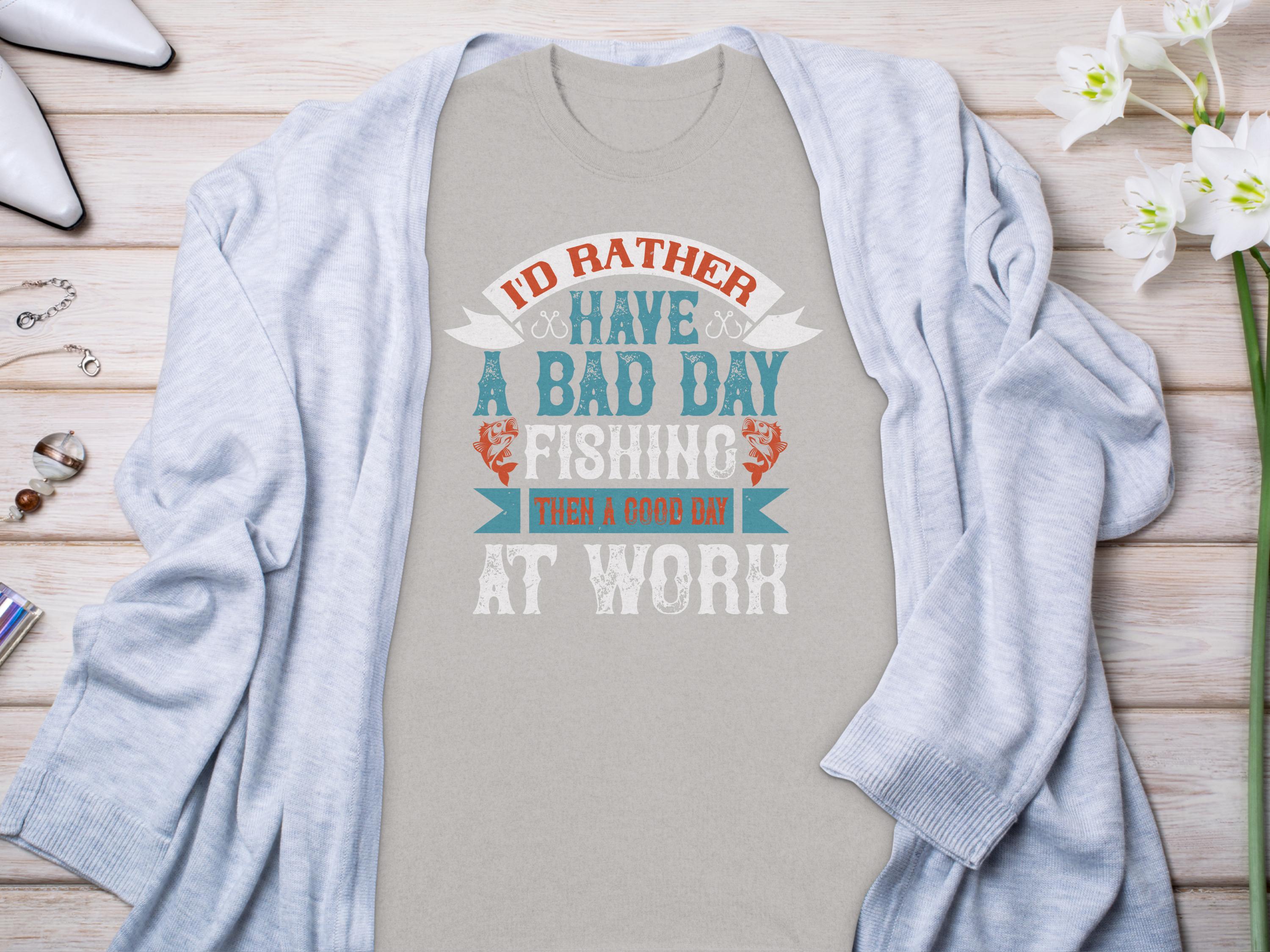 Displayed on a wooden surface, this Garment Graphics gray T-shirt, featuring Id rather have a bad day fishing than a good day at work, makes an ideal gift for anglers. Its partly covered by a light gray cardigan and accompanied by a flower, wristwatch, perfume, and nail polish.