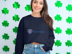 Wearing a black Garment Graphics Scania logo sweatshirt in unisex sizing and denim shorts, a smiling woman with long brown hair stands by a white wall decorated with green shamrocks, epitomizing the ideal casual outfit.