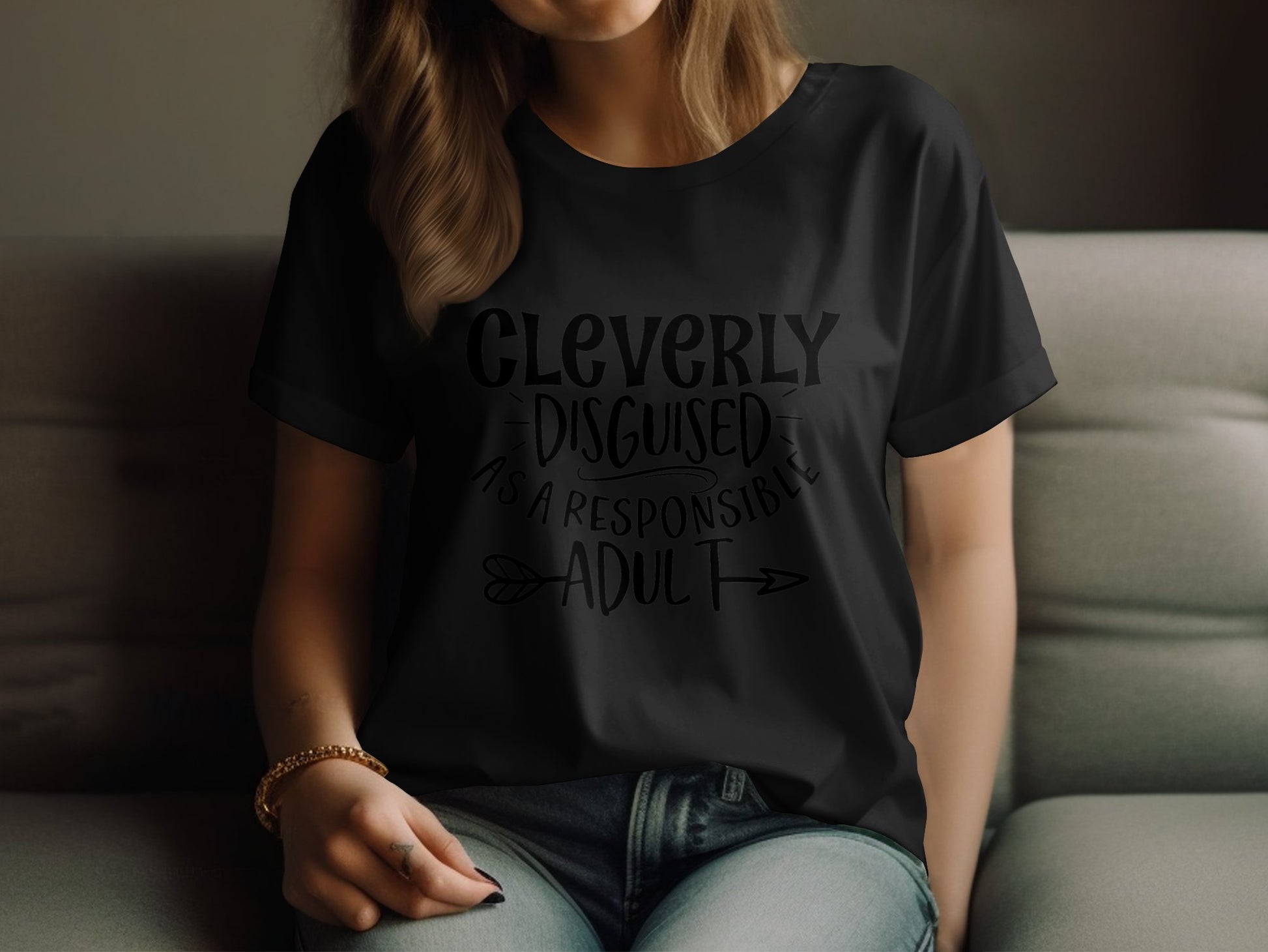 A person with light brown hair sits on a couch wearing a semi-fitted black t-shirt by Garment Graphics featuring the text Cleverly Disguised as a Responsible Adult, holding the edge of the shirt with their left hand.