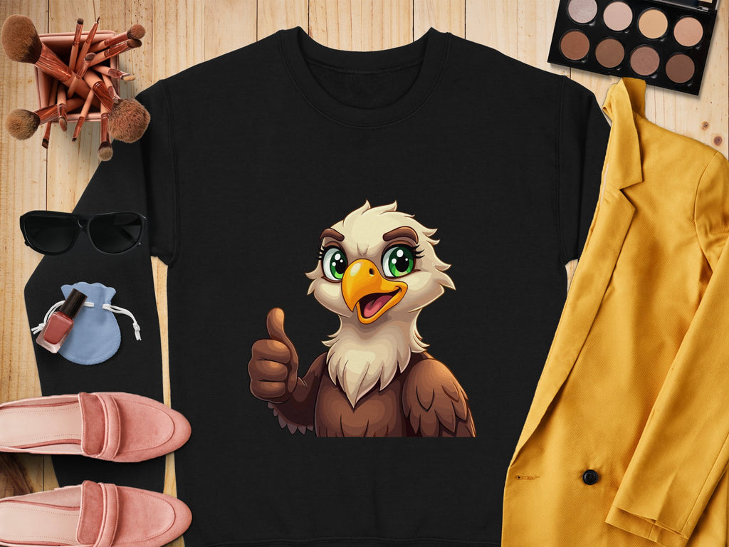 A Garment Graphics black shirt features a cheerful cartoon eagle giving a thumbs-up. The unisex shirt rests on wood, surrounded by makeup brushes, sunglasses, pink shoes, a yellow blazer, a blue pouch, lipstick, and an eyeshadow palette.