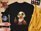A Garment Graphics black shirt features a cheerful cartoon eagle giving a thumbs-up. The unisex shirt rests on wood, surrounded by makeup brushes, sunglasses, pink shoes, a yellow blazer, a blue pouch, lipstick, and an eyeshadow palette.
