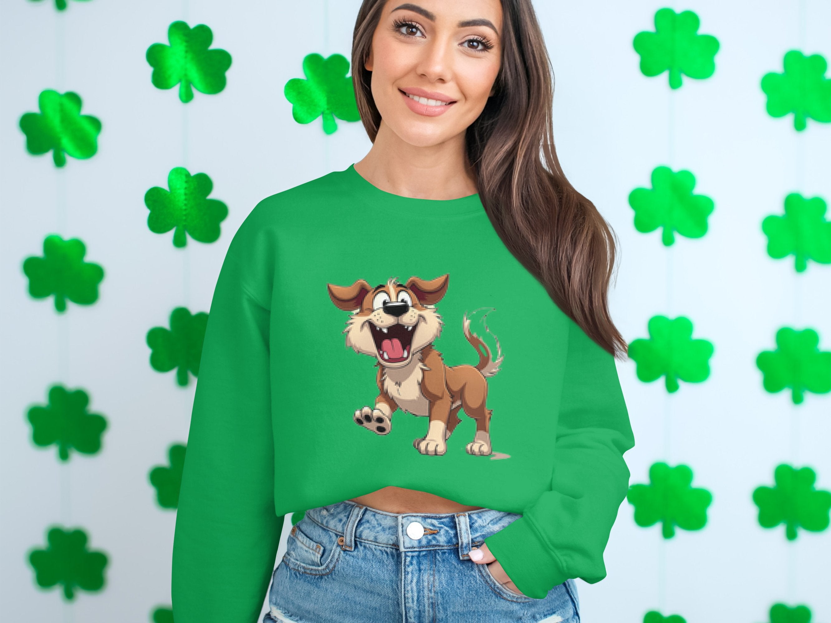 A person sports a green Garment Graphics sweatshirt featuring an animated happy dog illustration and denim shorts, smiling at the camera. The backdrop showcases a pattern of green shamrock cutouts, ideal for dog lovers on a vibrant day.