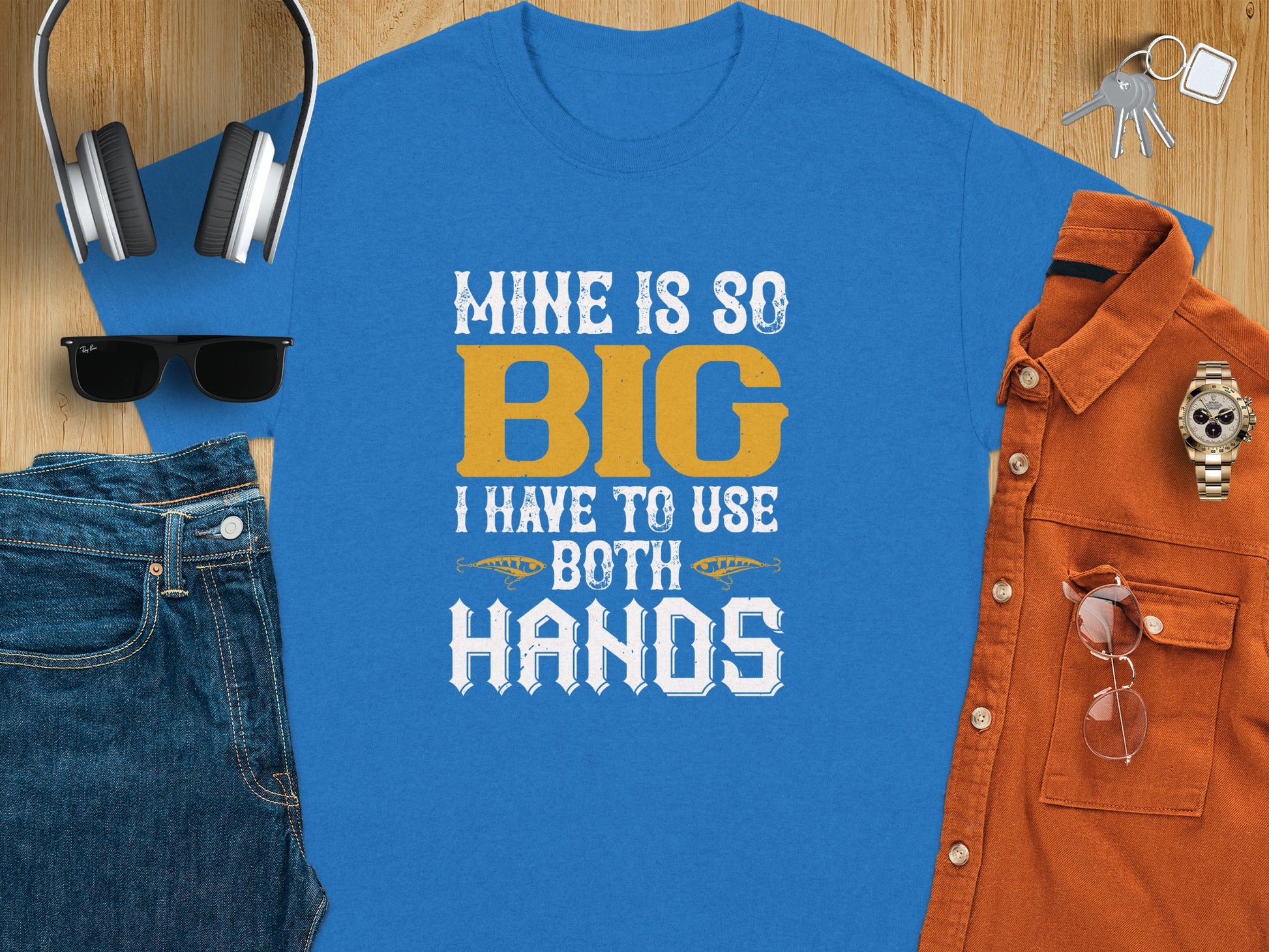 Garment Graphics presents a blue fishing-themed tee with the phrase Mine is so big I have to use both hands in white and yellow. Styled alongside headphones, a watch, sunglasses, jeans, keys, an orange shirt, and a notebook on wood—a humorous pick for fishing enthusiasts.