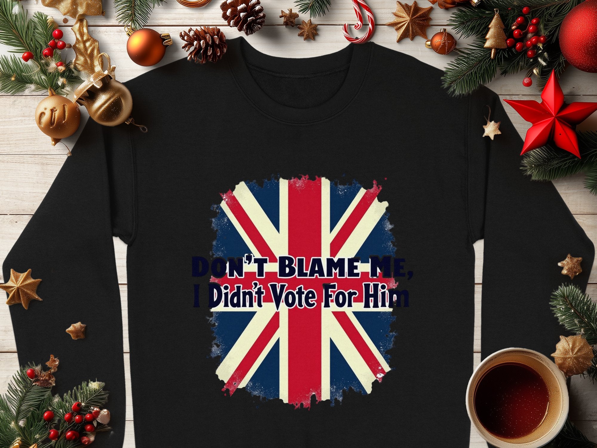 The black unisex sweatshirt by Garment Graphics, featuring a Union Jack and Dont Blame Me, I Didnt Vote For Him, is surrounded by festive Christmas décor like ornaments, pine branches, and a steaming mug.
