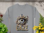 The Garment Graphics unisex graphic sweatshirt, displayed amid green plants and yellow flowers, showcases an ornate blue eye design adorned with jewels and pearls, with GAZE boldly printed below.