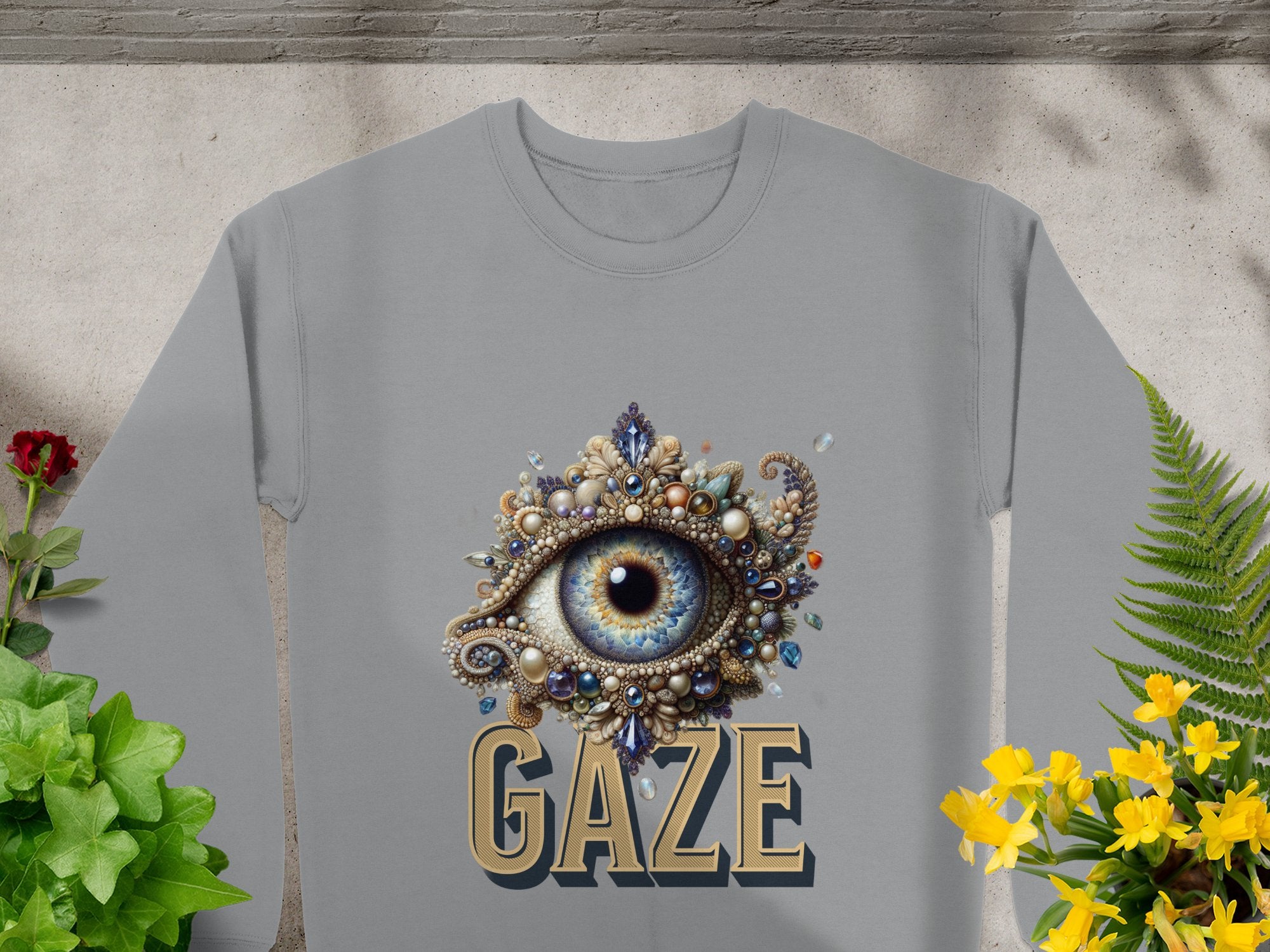 The Garment Graphics unisex graphic sweatshirt, displayed amid green plants and yellow flowers, showcases an ornate blue eye design adorned with jewels and pearls, with GAZE boldly printed below.