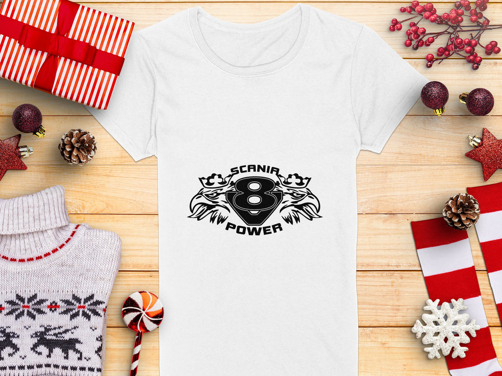 A white Garment Graphics T-shirt featuring a Scanir 8 Power logo with stylized helmets and abstract eagle heads is placed on a wooden surface surrounded by red and white wrapped gifts, pine cones, festive decorations, and a knitted sweater.