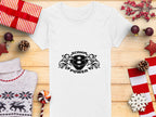 A white Garment Graphics T-shirt featuring a Scanir 8 Power logo with stylized helmets and abstract eagle heads is placed on a wooden surface surrounded by red and white wrapped gifts, pine cones, festive decorations, and a knitted sweater.