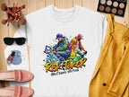 A white Garment Graphics t-shirt featuring two dancers with graffiti art and Risk Taker Street Dance 10th Final is displayed on a wooden surface. Its paired with a yellow blazer, pink loafers, black sunglasses, makeup essentials, a white watch, and a perfume bottle.
