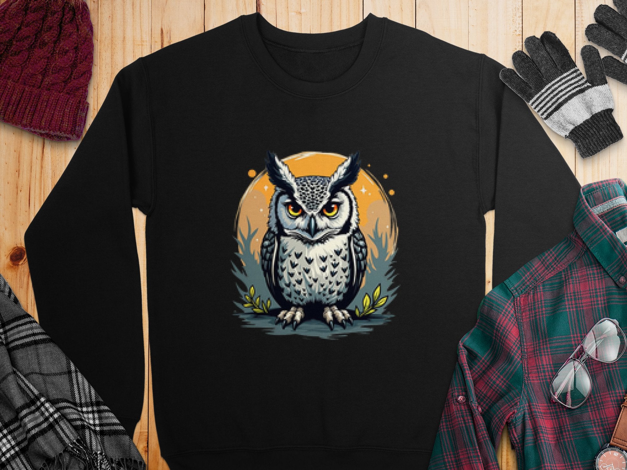 Garment Graphics black unisex sweatshirt features a stylized owl against an orange and starry backdrop. Its displayed on wood, paired with a red knit hat, plaid shirt, and gray gloves.