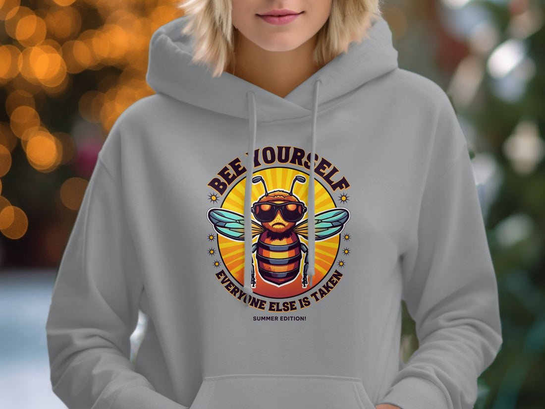 A white Bee Yourself Hoodie by Garment Graphics rests on a wooden surface, featuring a bee with sunglasses and the text Everyone Else is Taken, Summer Edition. Nearby are sandals, a hat, trendy sunglasses, a map-themed notebook, and a camera—ideal for making waves this summer.