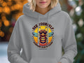 A person wears Garment Graphics gray Bee Yourself Hoodie, featuring an illustrated bee in sunglasses. The text says, Be Yourself, Everyone Else Is Taken, and Summer Edition, with vibrant blurred lights in the background.