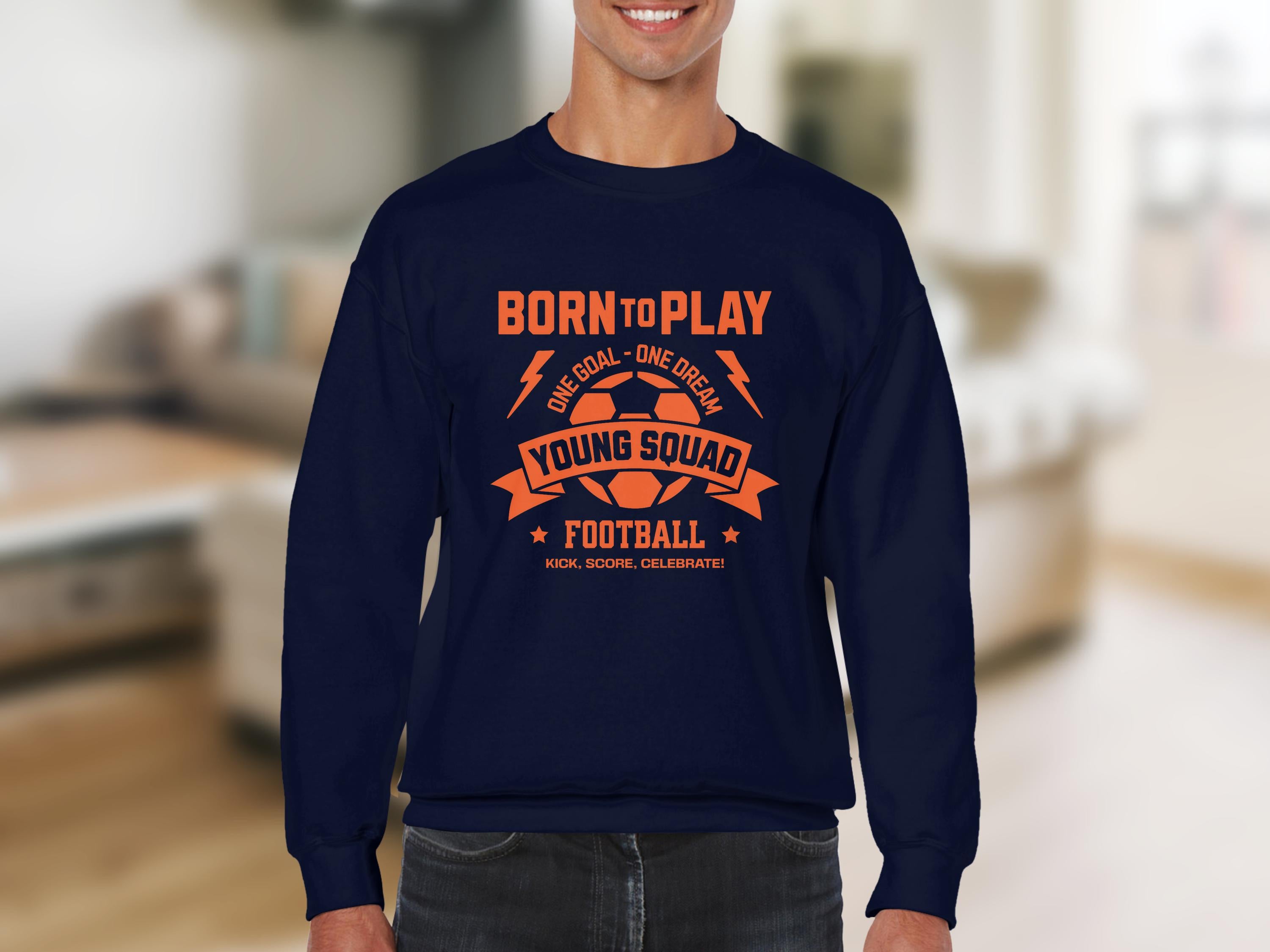 A person wears a navy Garment Graphics football sweatshirt with bold orange text: Born to Play, One Goal - One Dream, Young Squad, Football, and Kick, Score, Celebrate. Unisex sizing caters to young squads. The setting features a softly blurred interior room.
