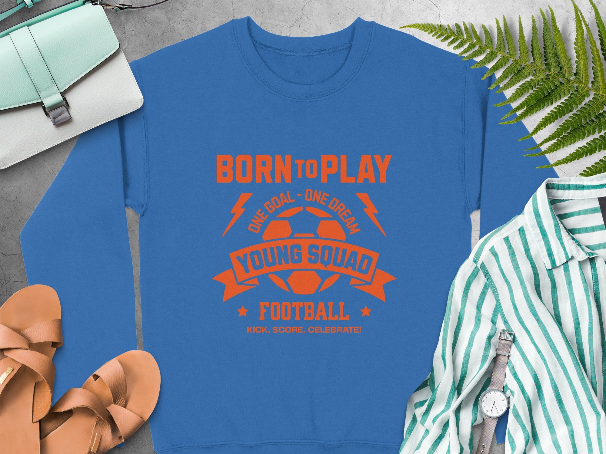 A blue football sweatshirt by Garment Graphics is displayed with an orange Born to Play and Young Squads Football soccer ball graphic. Nearby are a purse, sneakers, a striped shirt, and a fern leaf. It comes in unisex sizing for young squads.