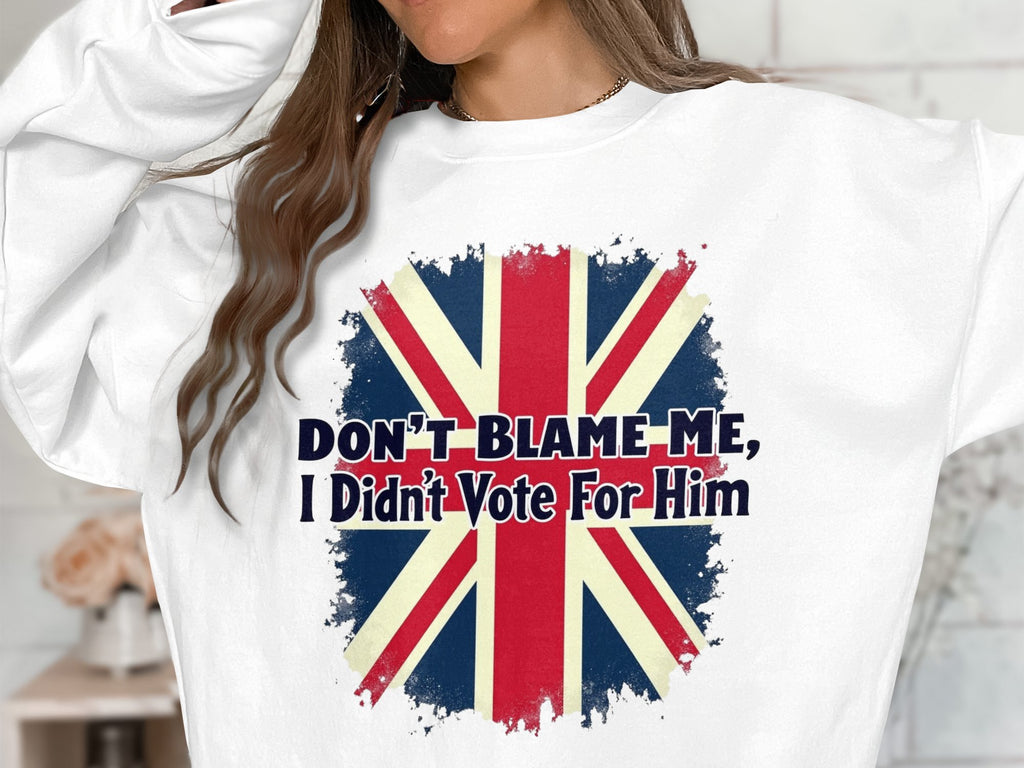 A person with long, wavy hair wears a Garment Graphics white unisex sweatshirt featuring the British flag and the text DONT BLAME ME, I Didnt Vote For Him. Their hair obscures their face.