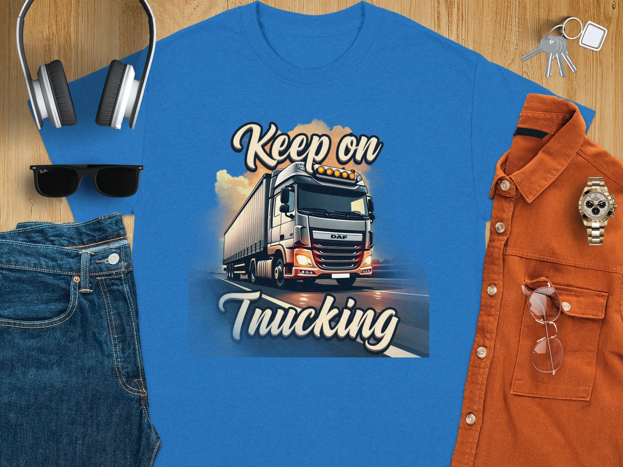 The Garment Graphics T-shirt boasts a blue graphic print with Keep on Trucking and a semi-truck illustration, ideal for truck enthusiasts. Paired with jeans, sunglasses, a watch, brown button-up shirt, and headphones on wood.