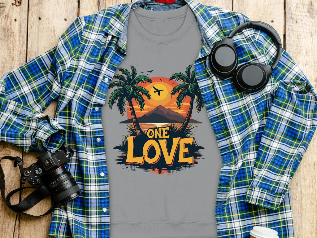 A flat lay presents a Garment Graphics gray T-shirt with a sunset, palm trees, and ONE LOVE, paired with a blue plaid shirt, headphones, camera, plant, and cup on wood. Ideal for One Love enthusiasts!.