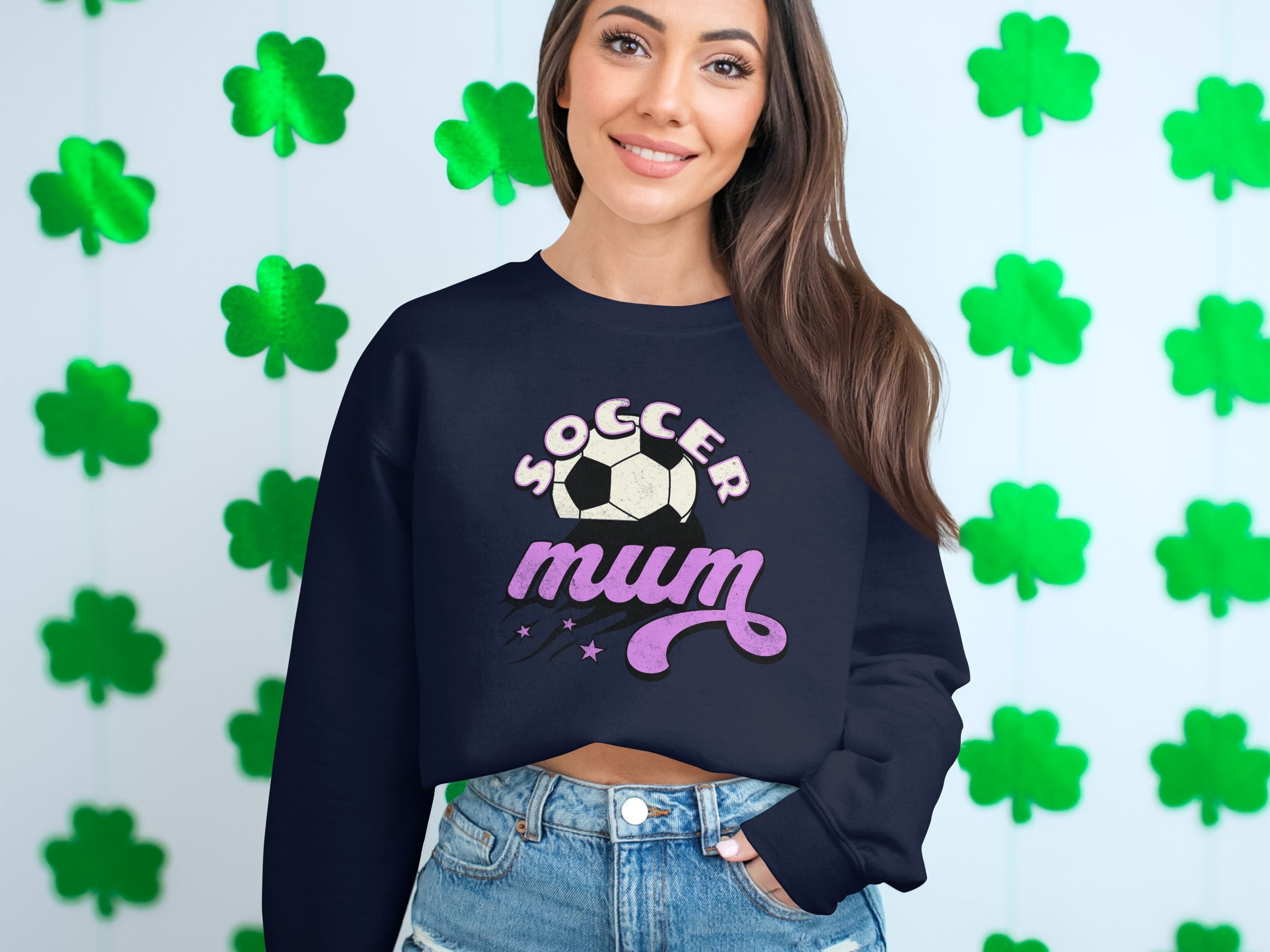 A woman wearing a Garment Graphics black sweatshirt with a vintage Soccer Mum graphic and soccer ball stands in front of a wall decorated with green shamrocks. She has long brown hair and is smiling.