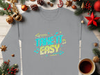 This gray Garment Graphics sweatshirt, featuring TAKE IT EASY in bold blue and yellow letters with arrows and musical notes, is showcased on a wooden surface surrounded by holiday decor and a steaming coffee cup, highlighting its trendy unisex design.