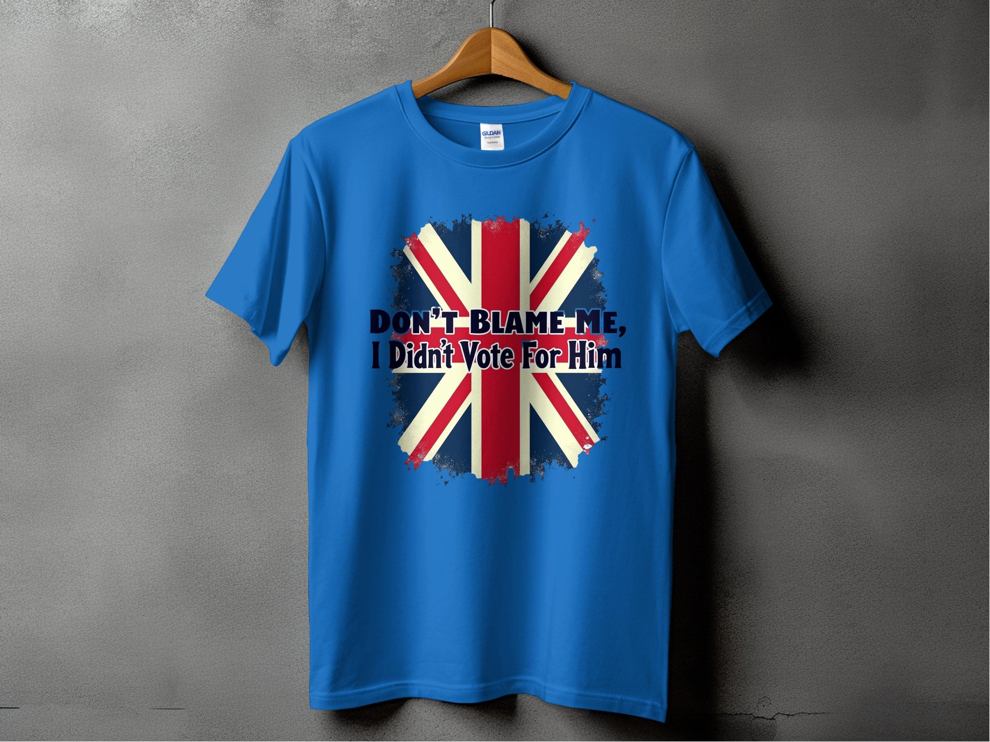 Garment Graphics presents a classic fit blue T-shirt on a wooden hanger, featuring a bold UK flag graphic with the statement DONT BLAME ME, I Didnt Vote for Him against a dark gray backdrop.