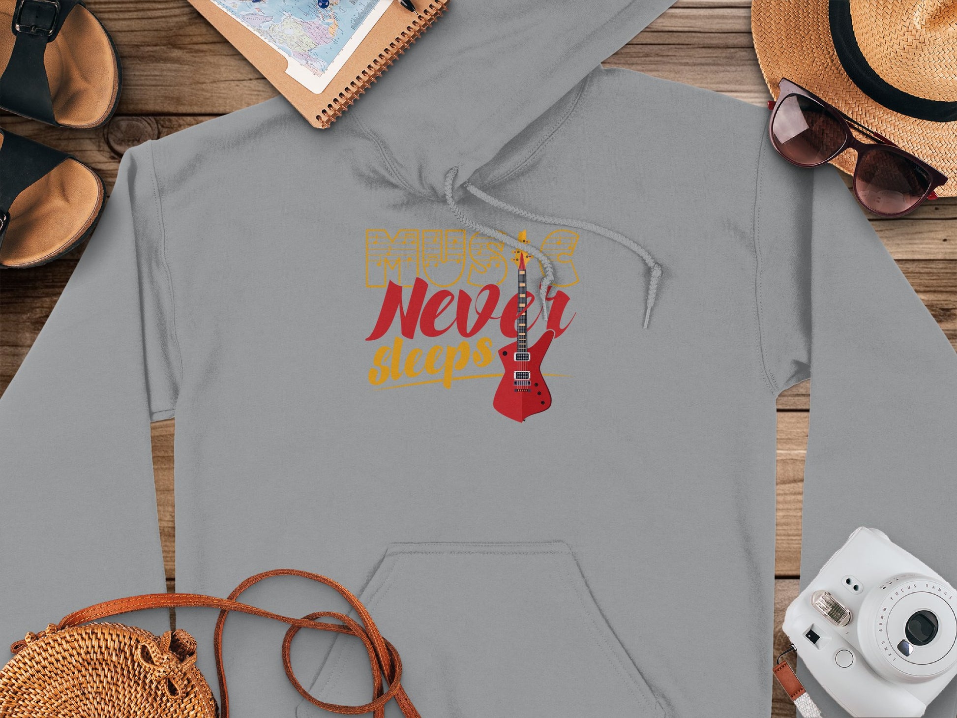 The Garment Graphics gray hoodie has a classic fit, featuring Music Never Sleeps and a red electric guitar graphic. Its surrounded by a map, sunglasses, straw hat, sandals, camera, and woven bag on wood.