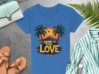The Garment Graphics blue t-shirt features a tropical sunset with palm trees, a soaring bird, mountains, and One Love text. Ideal for beach outings, pair it with sandals, a green striped shirt, watch, and purse on the gray surface.