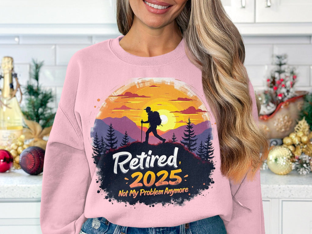 A person wears a Garment Graphics pink retirement sweatshirt with a silhouetted hiker against a sunset, featuring the text Retired 2025, Not My Problem Anymore. The unisex sweatshirt is showcased in a festively decorated white kitchen.