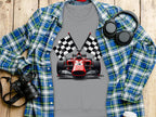 A Garment Graphics gray t-shirt with a racing car and checkered flags is displayed on wood, ideal for motorsport fans. Its paired with a blue and green plaid shirt. Black headphones, a camera, and a white cup round out the scene.