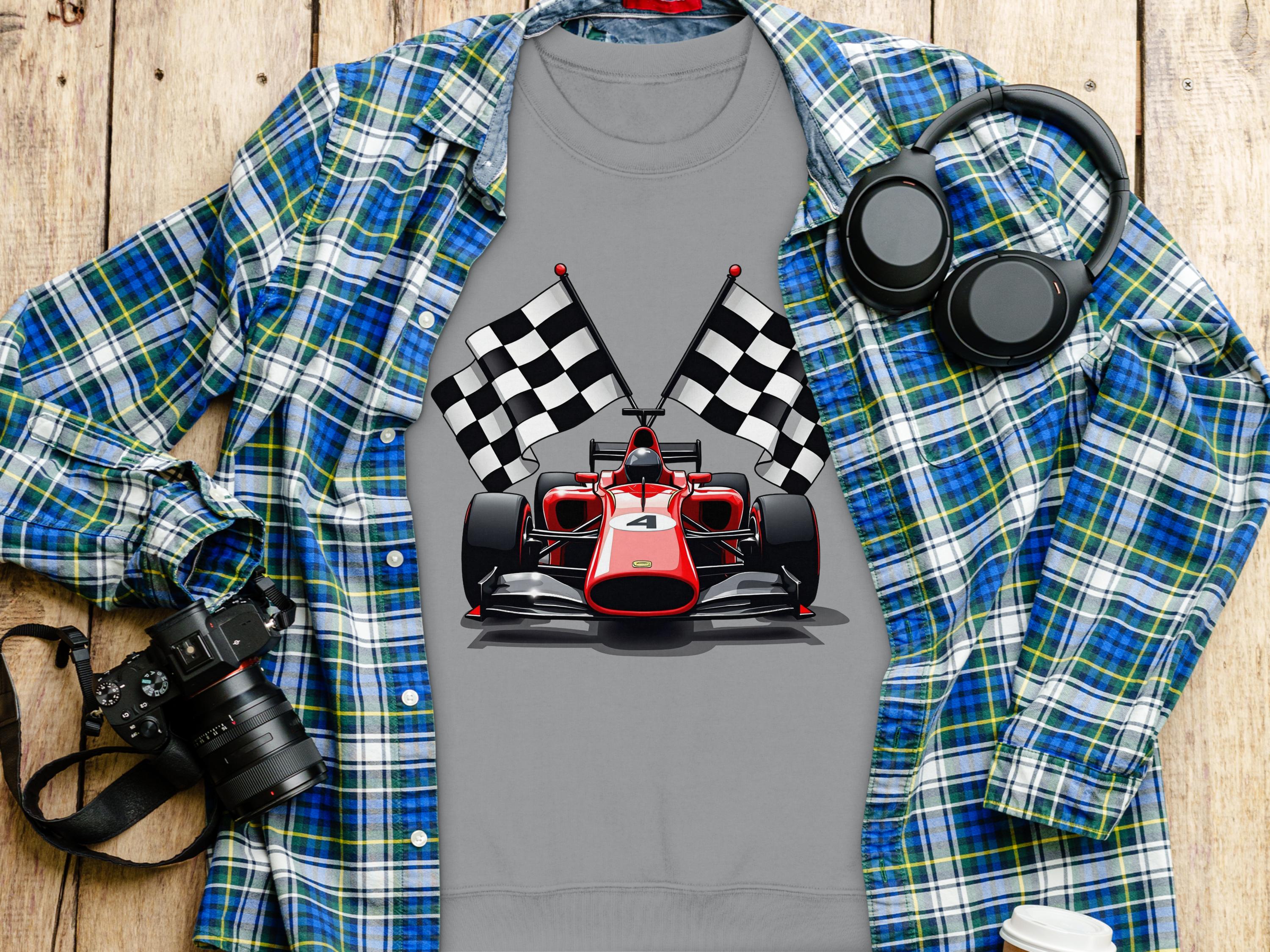 A Garment Graphics gray t-shirt with a racing car and checkered flags is displayed on wood, ideal for motorsport fans. Its paired with a blue and green plaid shirt. Black headphones, a camera, and a white cup round out the scene.