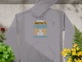 The Garment Graphics gray hoodie features the design Im not addicted to fishing, we are just in a very committed relationship in white, yellow, and blue fonts. This classic-fit fishing hoodie is showcased on a surface surrounded by potted plants and flowers.