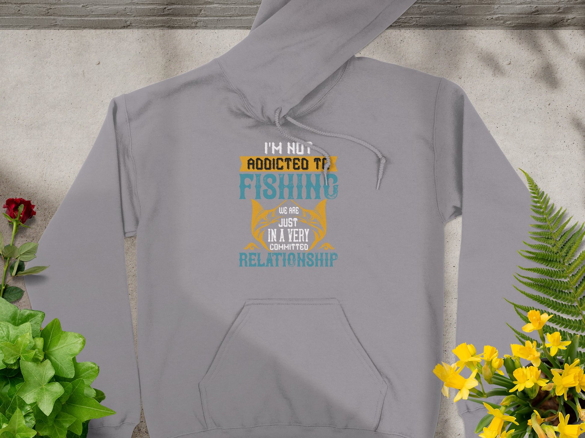 The Garment Graphics gray hoodie features the design Im not addicted to fishing, we are just in a very committed relationship in white, yellow, and blue fonts. This classic-fit fishing hoodie is showcased on a surface surrounded by potted plants and flowers.