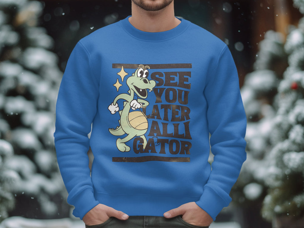 A person wears a blue graphic sweatshirt from Garment Graphics featuring a cartoon alligator dribbling a basketball with the text See You Later Alligator, set against blurred, snow-covered trees.