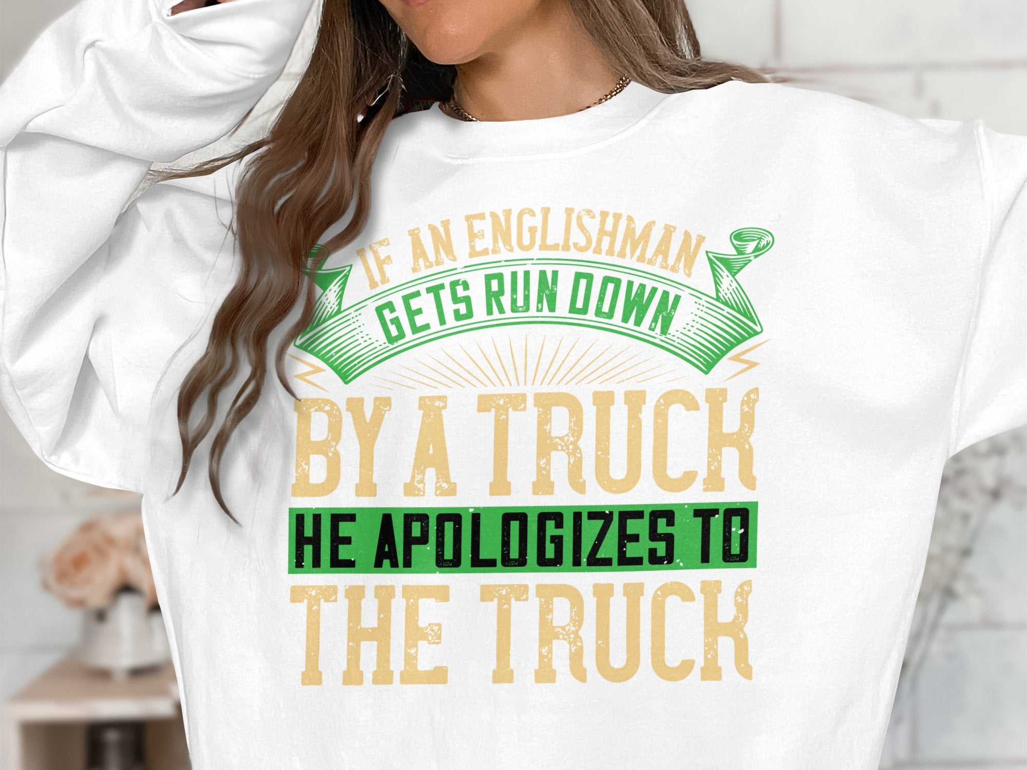 A person wears a white Garment Graphics sweatshirt featuring a humorous graphic with the words If an Englishman gets run down by a truck, he apologizes to the truck in green, yellow, and tan. Available in unisex sizes.